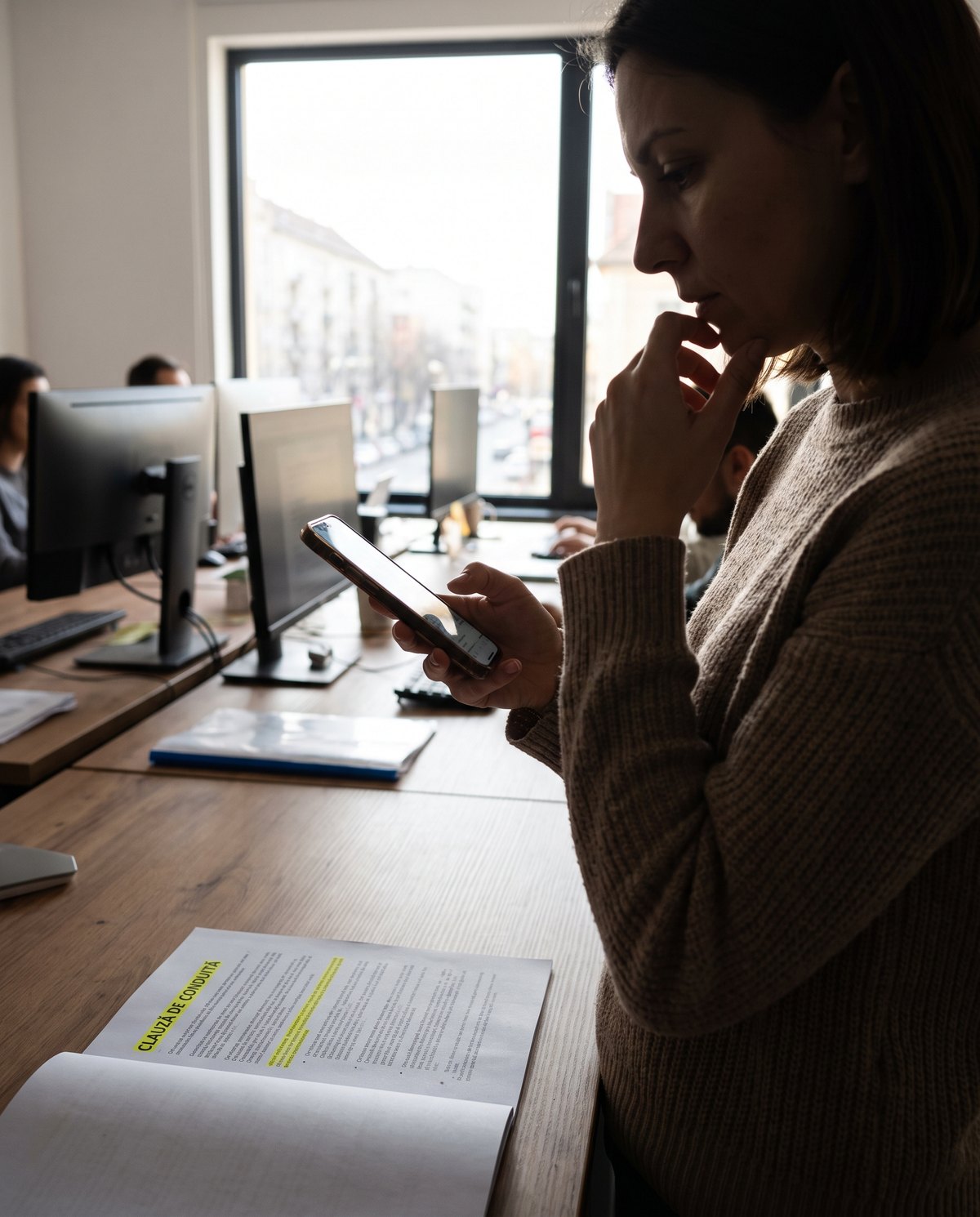 O femeie stând lângă un birou într-un birou, privindu-și telefonul cu gândul, cu o mână pe bărbie; în prim-plan se vede un caiet deschis cu text subliniat, iar în fundal se văd oameni care lucrează la calculatoare.