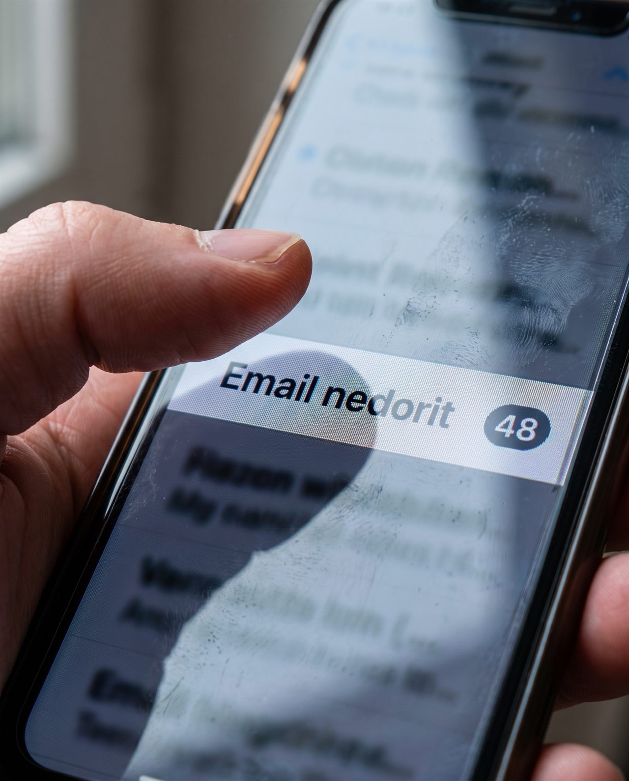 Un deget atinge ecranul unui smartphone pe care se afișează o aplicație de e-mail cu un dosar evidențiat, intitulat „Email nedorit”, și „48” de mesaje necitite. Pe ecran se văd pete și reflexii.