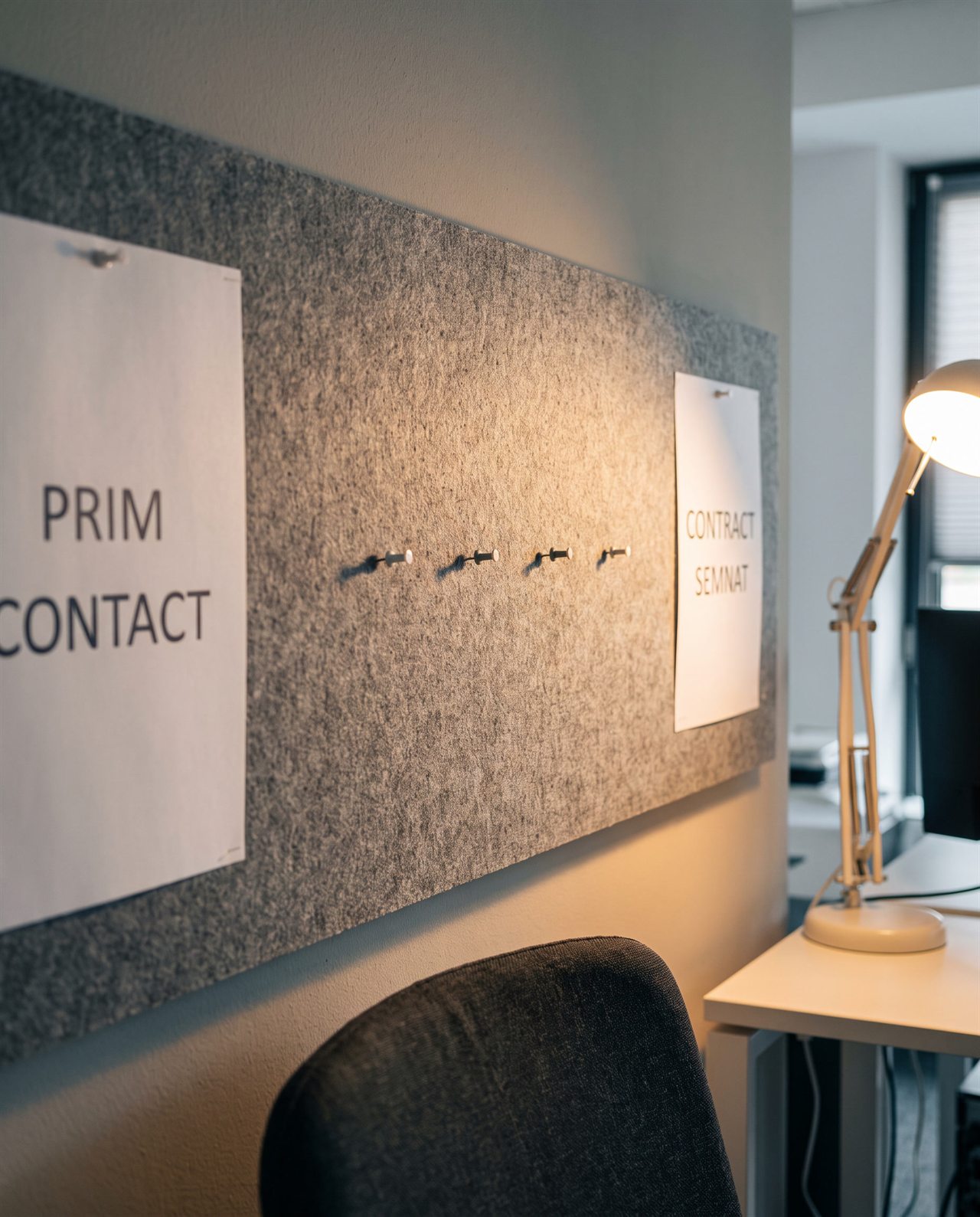 Un perete modern de birou, pe care sunt fixate cu pioneze două foi de hârtie pe o tablă de fetru gri, cu inscripțiile „PRIM CONTACT” și „CONTRACT SEMINAT”. În apropiere se văd o lampă și o parte a unui birou cu un monitor.