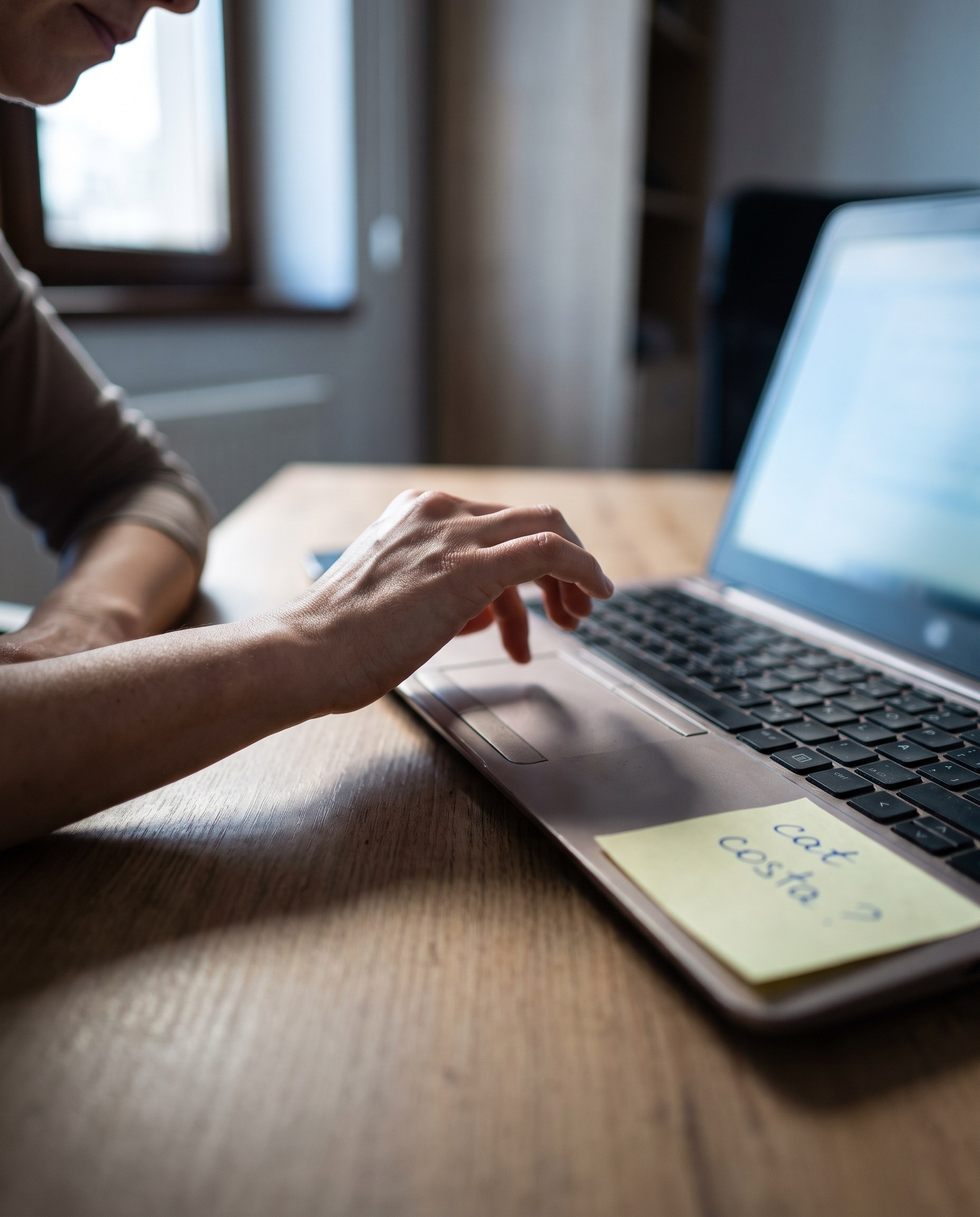 Mână oprită deasupra trackpadului unui laptop, lângă un bilețel galben pe care scrie cât costă